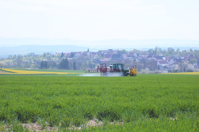 Das Bild zeigt ein landwirtschaftliches Gerät, mit dem vor dem Hintergrund eines Dorfes in Deutschland ein Feld intensiv bewirtschaftet wird